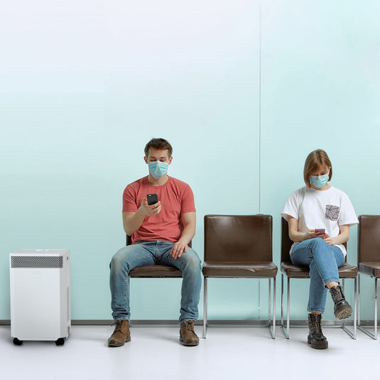 A waiting room at a doctor's surgery. Two people are sitting on chairs waiting for their appointments. There is an INOVA air purifier to the side, quietly cleaning the air, reducing the risk of viral infection.